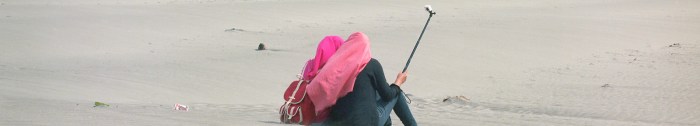 A photograph of the backs of two young Indonesian women, both wearing head scarves, and wielding a camera on a selfie stick